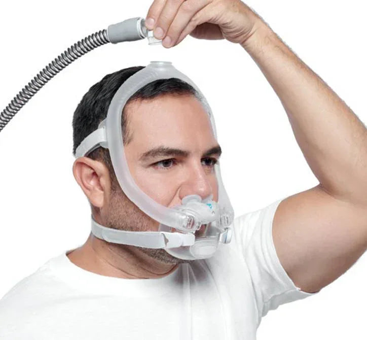 Man wearing a full face CPAP mask with headgear, adjusting the connected breathing hose