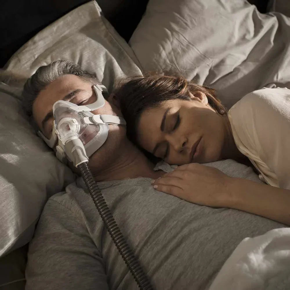 Man sleeping with CPAP mask and hose for sleep apnea therapy, woman resting beside him