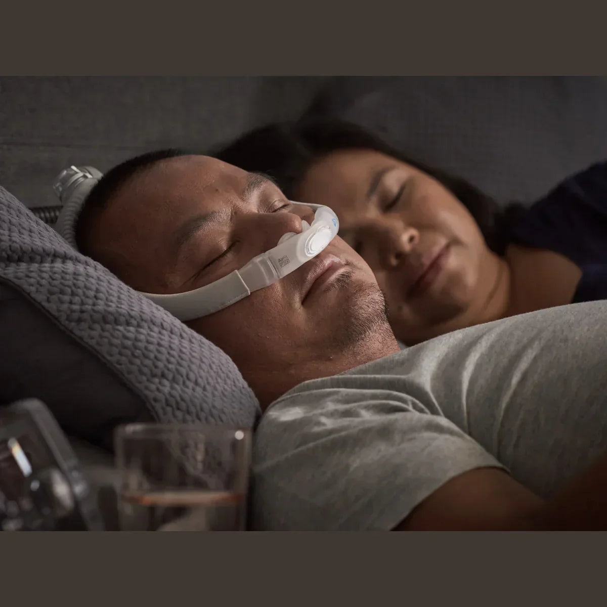 Man sleeping on bed wearing nasal pillow CPAP mask, woman sleeping beside him