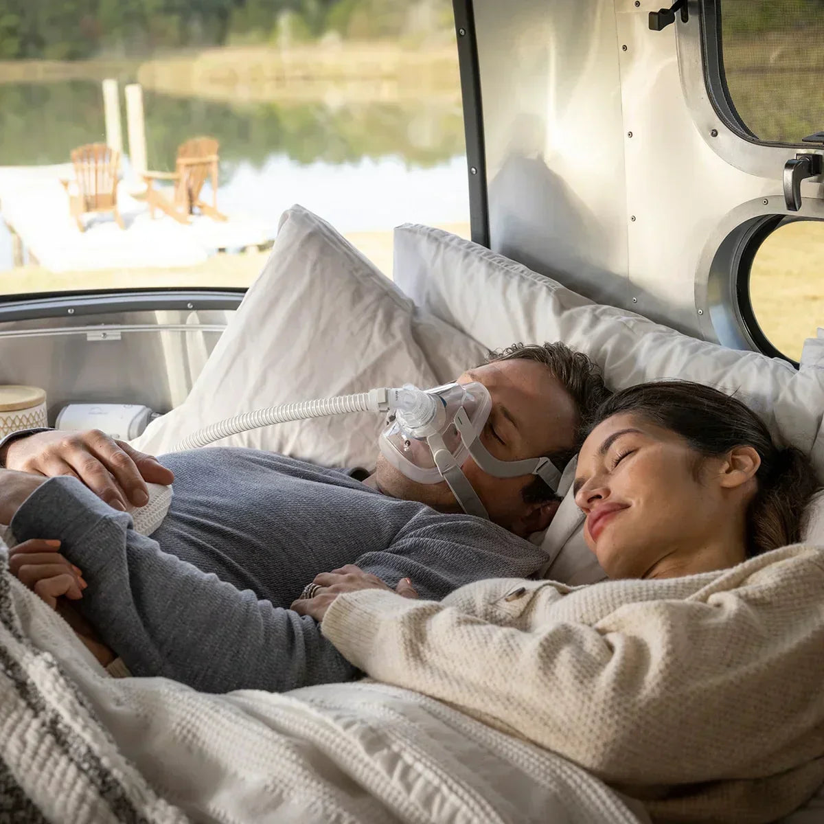 Man sleeping with CPAP mask and tube in bed next to woman, portable CPAP machine in background