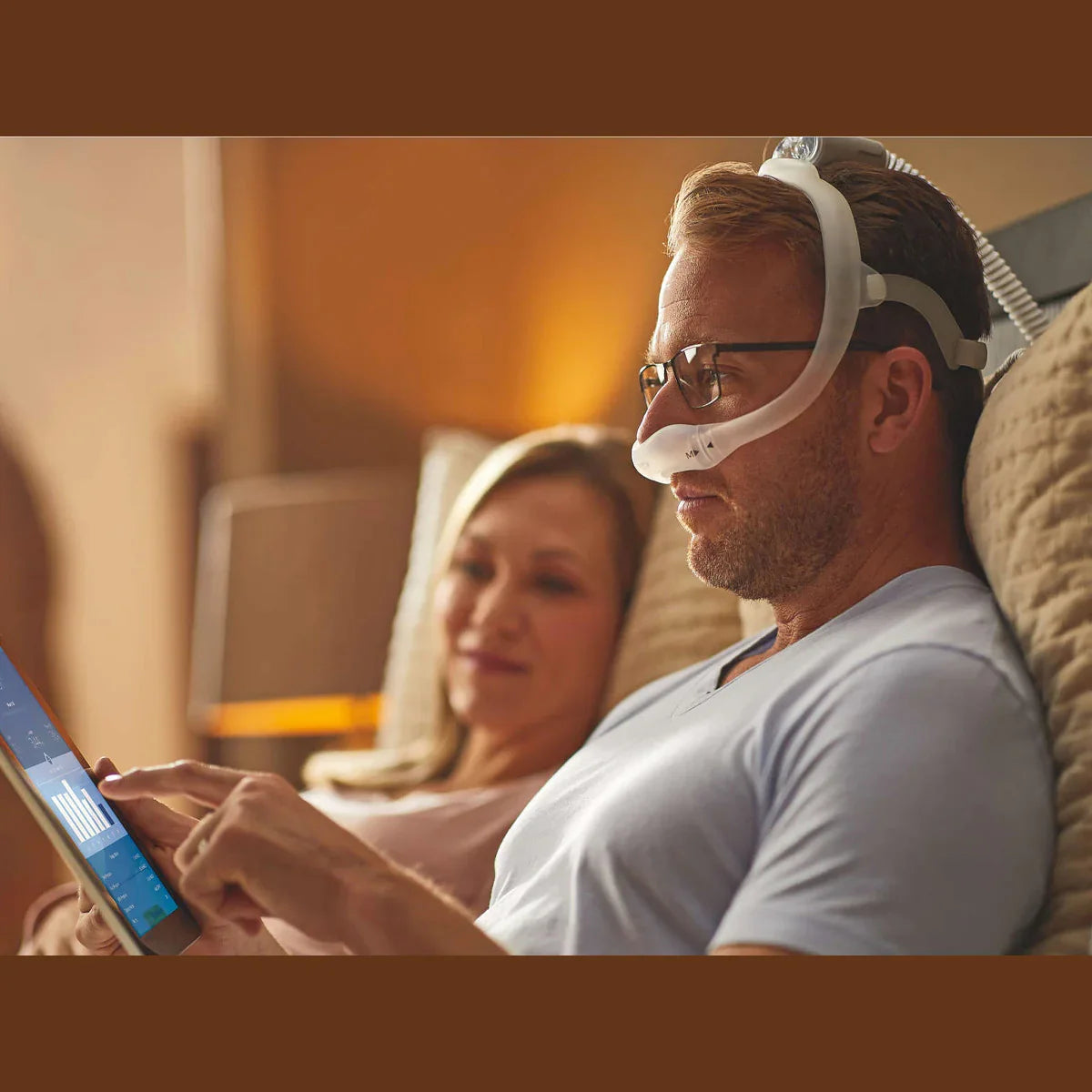 Man wearing a nasal CPAP mask using a tablet in bed, woman relaxing beside him