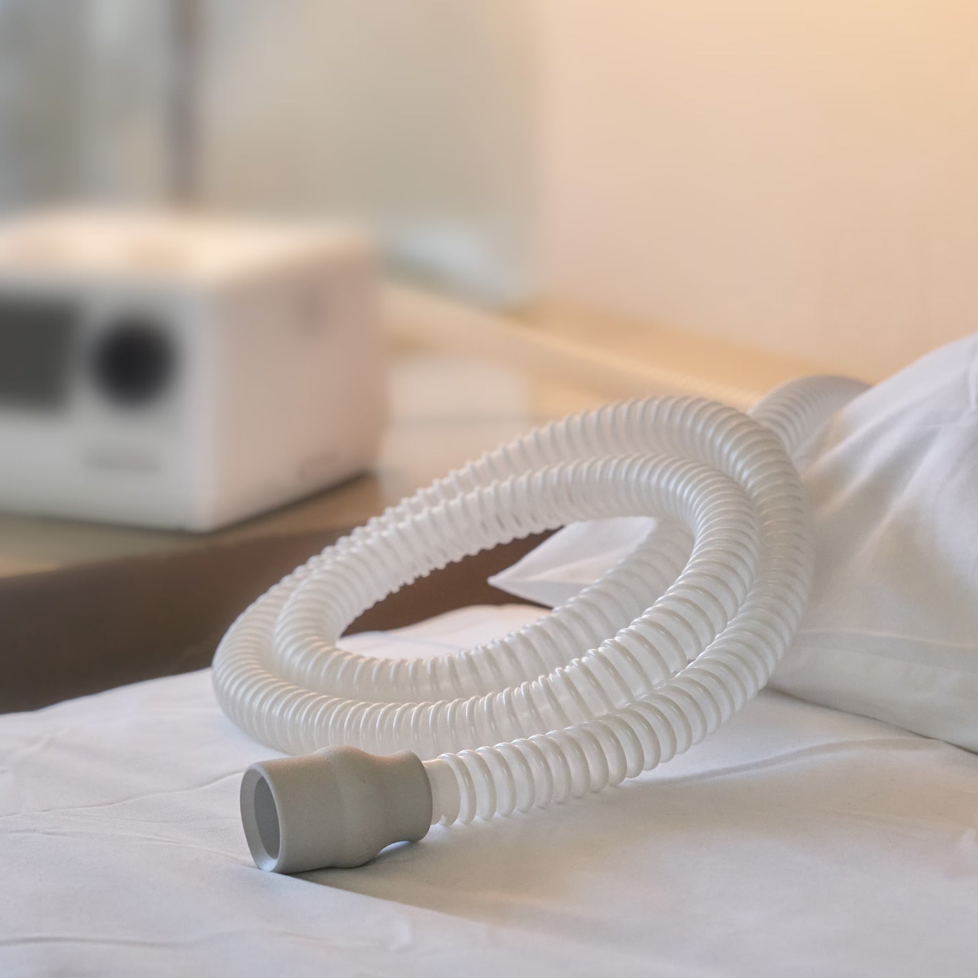 A white corrugated CPAP tube with a gray connector resting on white bedding, with a blurred medical device in the background.