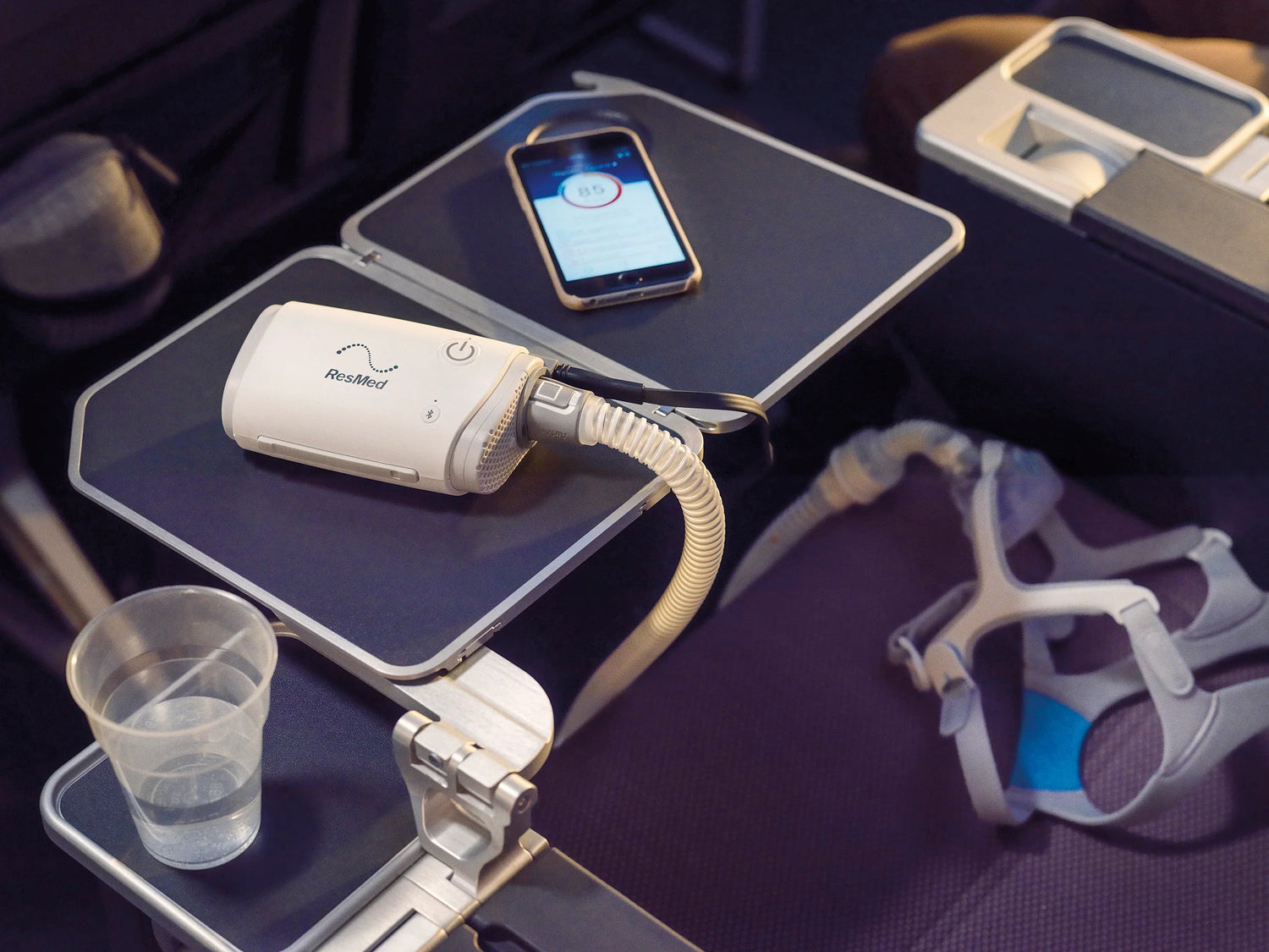 A portable medical oxygen concentrator with a curved hose and face mask resting on a dark airplane tray table, alongside a smartphone, a clear plastic cup, and an oxygen face mask on a purple seat.