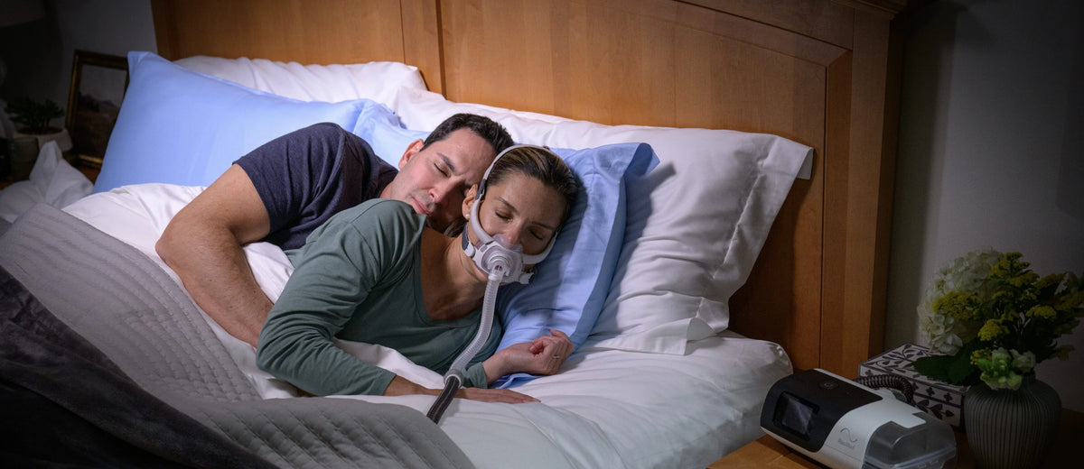 A couple lying in bed wearing a CPAP Mask, with a sleep apnea device on a bedside table in a dimly lit bedroom.